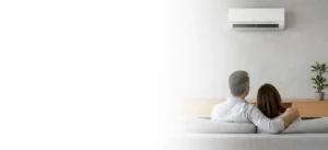 Couple sitting on a light gray sofa, facing a wall-mounted air conditioner in a modern living room.