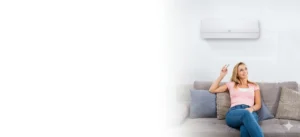 Woman sitting on a gray sofa in a living room, holding a remote and looking at a wall-mounted air conditioner above her.