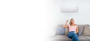 Woman relaxing on a sofa in a living room, holding a remote while looking up at a wall-mounted air conditioner above her.