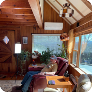 Man in a beige shirt and cap relaxes in a burgundy leather recliner in a sunlit, wood-paneled living room with plants and large windows.