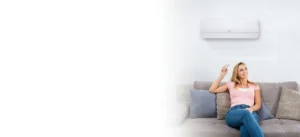 Woman relaxing on a gray couch, holding a remote and looking up at a wall-mounted air conditioner.