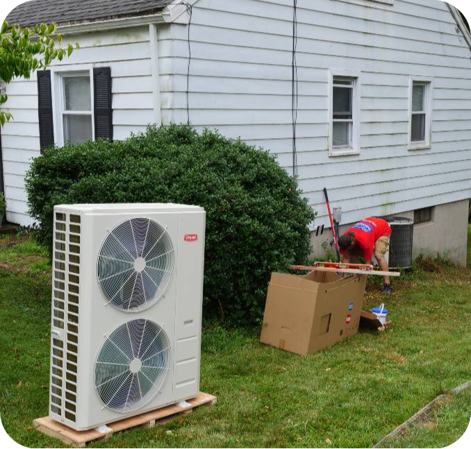 Outdoor air conditioner condenser on a wooden pallet in a grassy yard beside a large shrub, with a person in a red shirt working near cardboard boxes nearby.