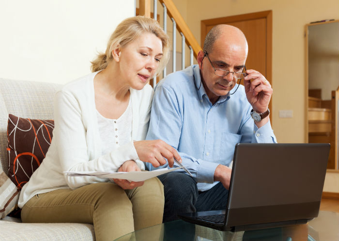 Couple sits on a couch looking at a laptop and papers together, appears to be reviewing finances or documents at home.